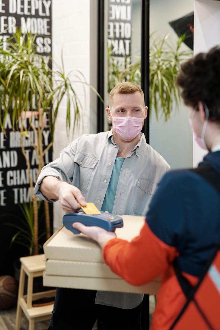 Man wearing face mask processing contactless payment for food delivery indoors.